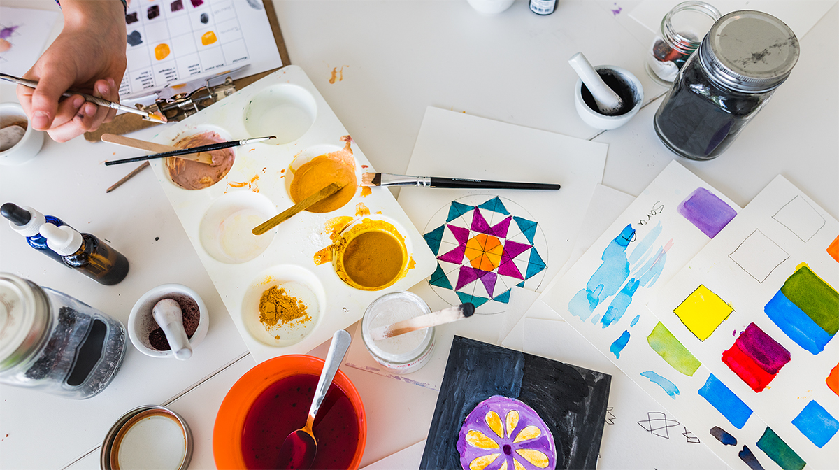 Paintings and paintbrushes are spread across a table with a child’s hand reaching for paint.